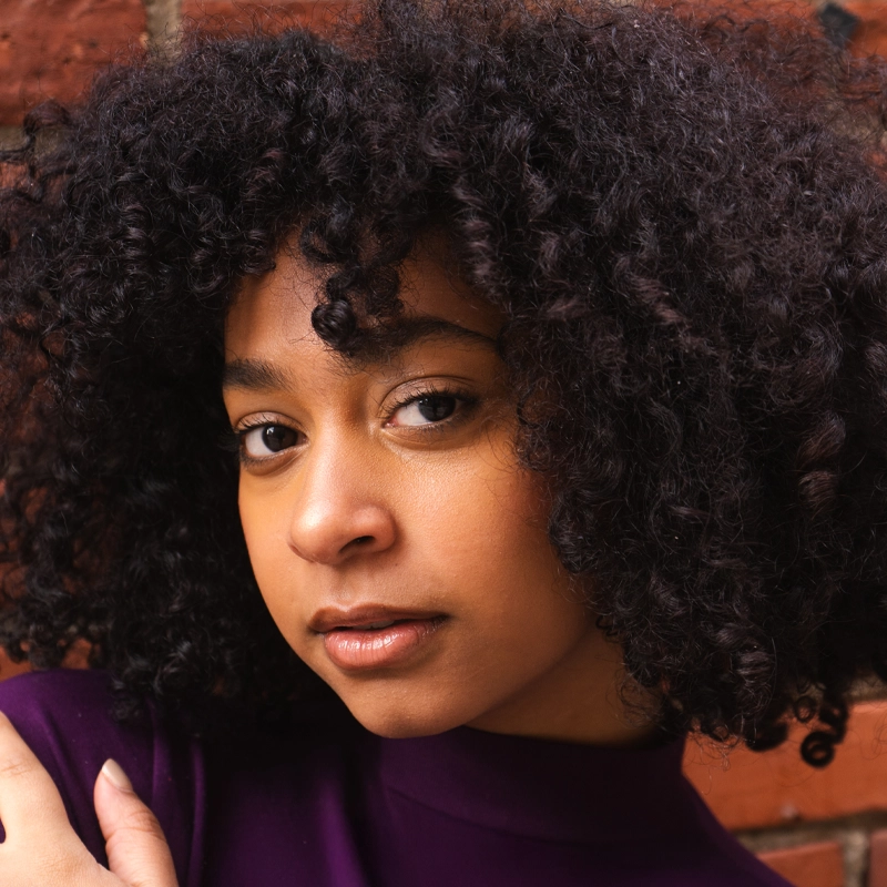 A young person with medium-dark skin and natural, curly black hair looks softly at the camera. Brianna Johnson wears a purple top and stands against a textured red brick wall. The lighting highlights her smooth complexion and relaxed expression.