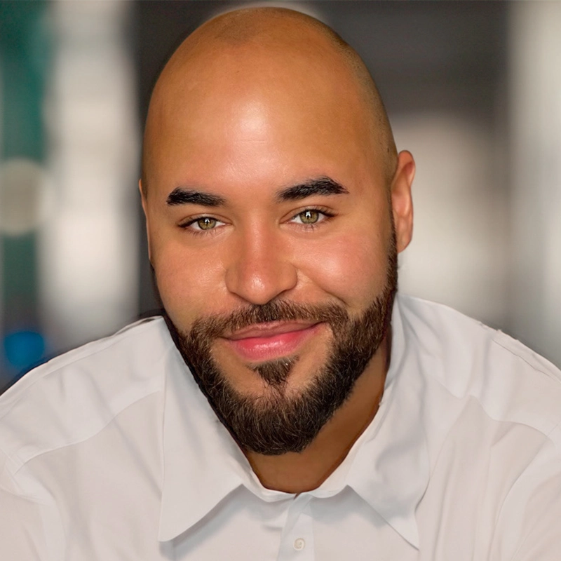 A bald man with a trimmed beard and mustache smiles slightly. He has light eyes, shaped eyebrows, and wears a white collared shirt. The blurred background is neutral, featuring soft hues of blue and gray. Lighting highlights his face.