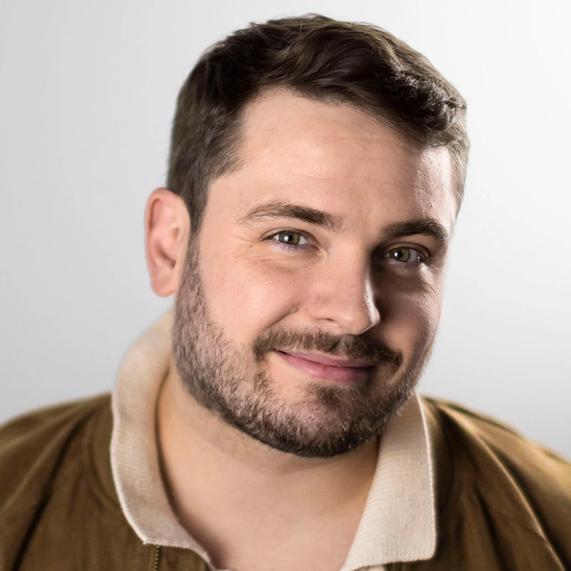 A man with short brown hair, trimmed beard, and light eyes smiles gently at the camera. He wears a tan jacket with a cream collar over a white shirt. The background is simple and light gray, giving a soft and friendly portrait feel.