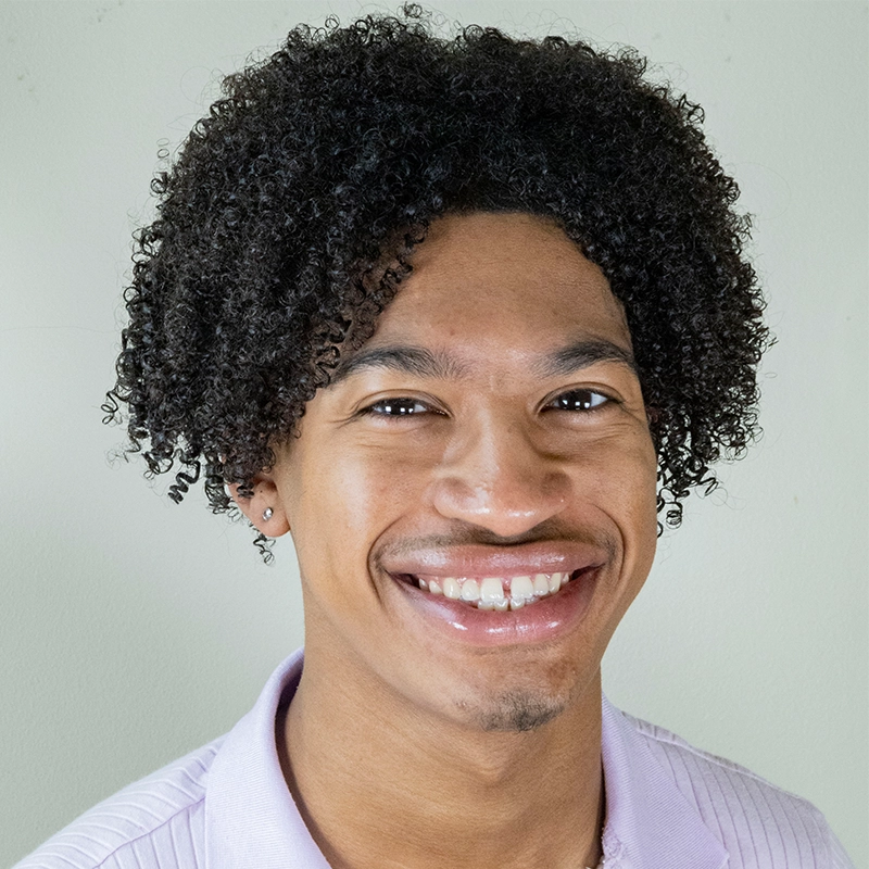 A young person with medium-brown skin and curly, dark hair smiles at the camera. Phillip Wong wears a light lavender collared shirt and a small earring in their left ear. The background is plain and light-colored.