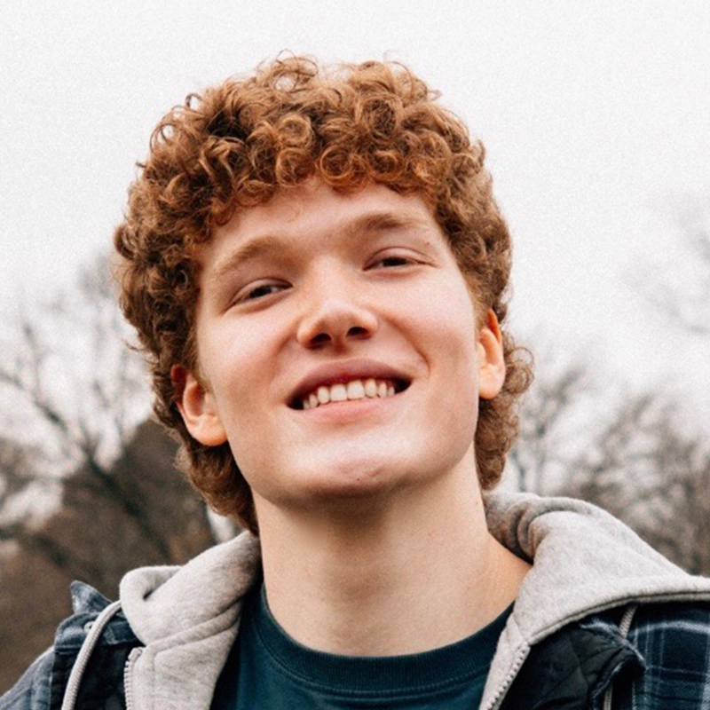 Jules Kessler, a young person with short, curly reddish-brown hair, smiles outdoors. They wear a dark jacket layered over a gray hoodie and a teal shirt. Leafless trees and an overcast sky fill the background.