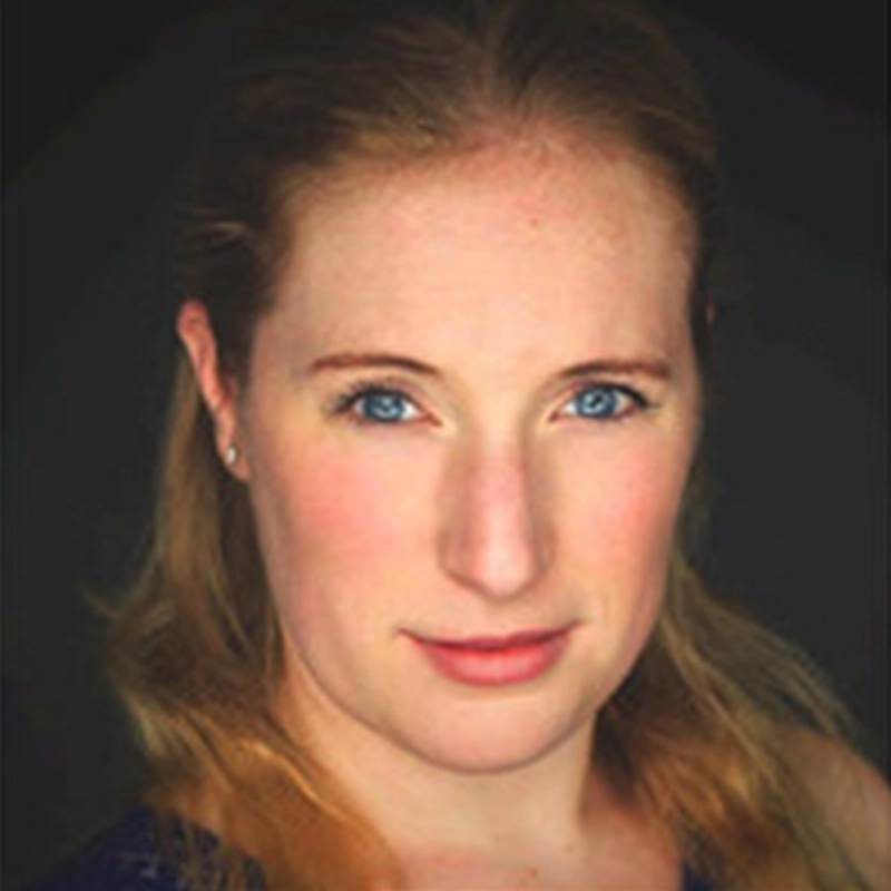 A woman with long, light brown hair and blue eyes looks directly at the camera. She has fair skin, subtle makeup, and wears small earrings. The background is dark—an elegant setting often favored by photographers like Patrick Pevehouse—making her face the primary focus of the image.