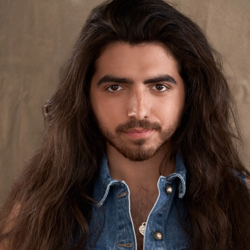 A person with long dark hair, a beard, and mustache stares directly at the camera. They wear a sleeveless blue denim shirt with buttons. The background is plain brown and softly lit, giving a warm tone to the image. The subject exudes quiet confidence, reminiscent of Charles Kadair’s distinctive style.