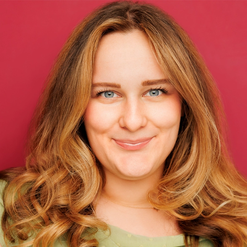 Jayline Ross, a woman with long, wavy blonde hair and blue eyes, smiles gently at the camera. She is wearing a light green top against a solid deep pink background. Bright lighting highlights her facial features and hair.