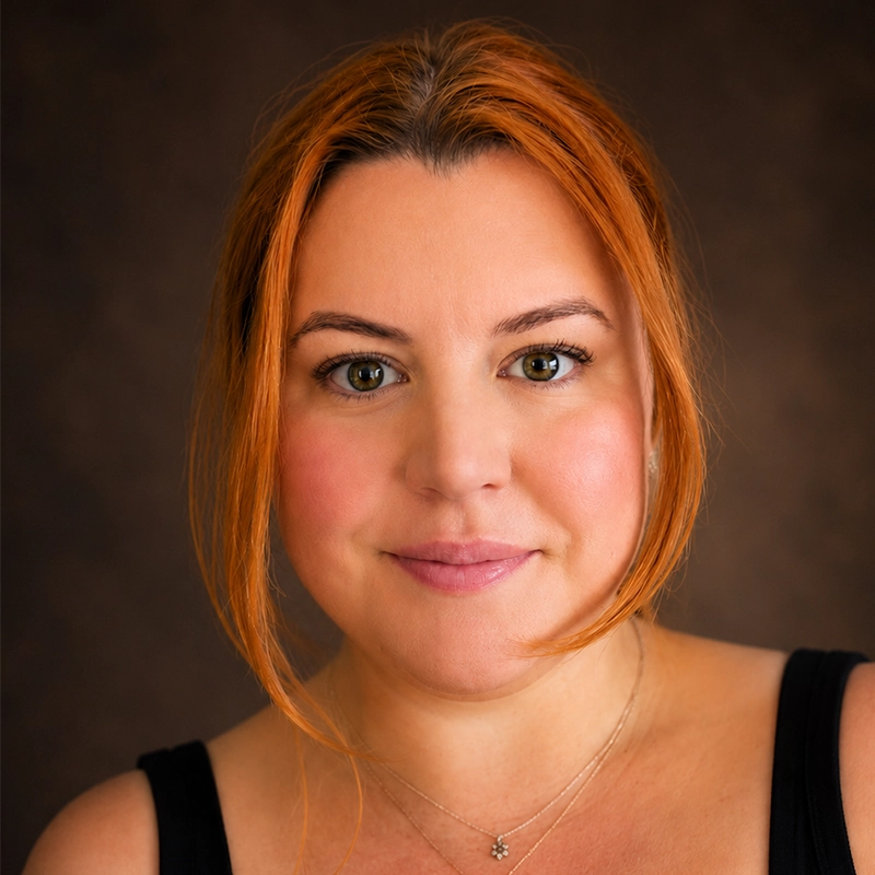 A woman with fair skin and long reddish-orange hair parted in the middle wears a black tank top and a delicate necklace with a small pendant. She is smiling softly, looking directly at the camera, against a dark brown, softly blurred background.