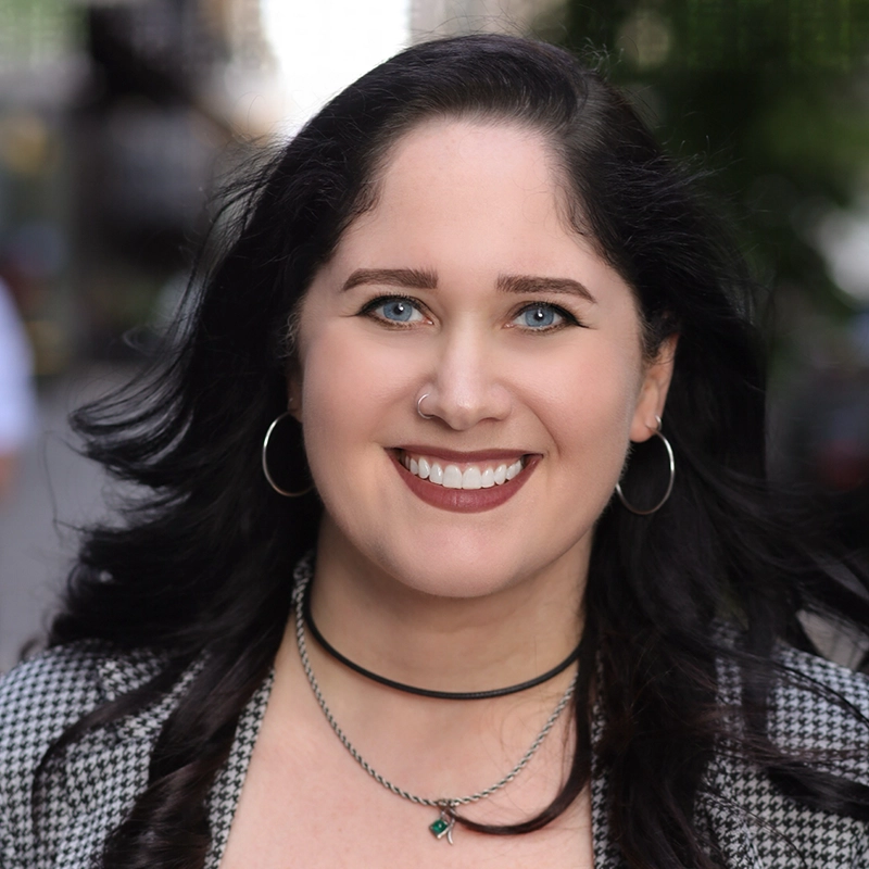 Alexandra Zografos, a woman with long dark hair, blue eyes, and light skin, smiles outdoors. She wears hoop earrings, two necklaces, a nose ring, and a black-and-white checkered jacket. The background is blurred city buildings and greenery.
