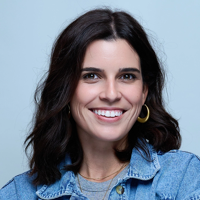 Kaylie Ann Delauri, with shoulder-length dark brown hair, gold hoop earrings, layered necklaces, and a denim jacket, smiles broadly against a plain light blue background.