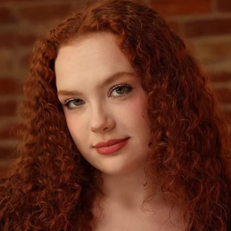A woman with long, curly red hair and fair skin looks at the camera with a soft smile. She has light makeup and is posed in front of a blurred brick wall background.