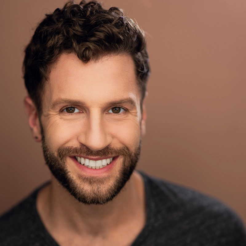 Zachary Kropp, a man with curly brown hair and a trimmed beard, smiles broadly. He has light skin, wears a dark V-neck shirt, and has a small earring in his left ear. The background is a soft, warm brown, creating a friendly, inviting portrait.
