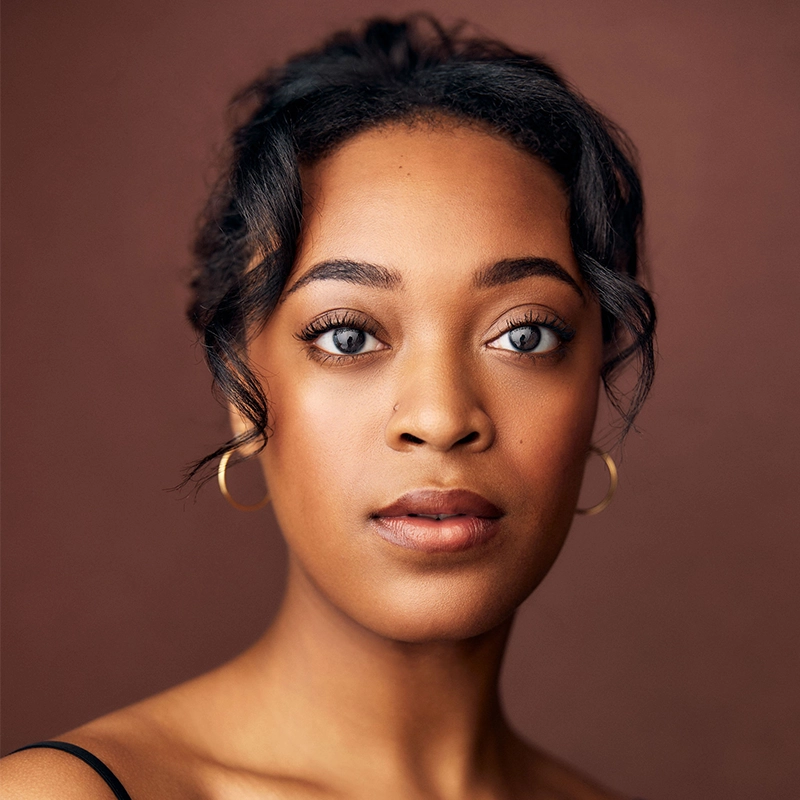 A woman with medium-brown skin, wavy dark hair pulled back, and gold hoop earrings looks directly at the camera. She has soft makeup, full eyebrows, and wears a thin-strap top. The portrait captures the understated elegance reminiscent of Asher Alcantara’s style against a plain brown background.