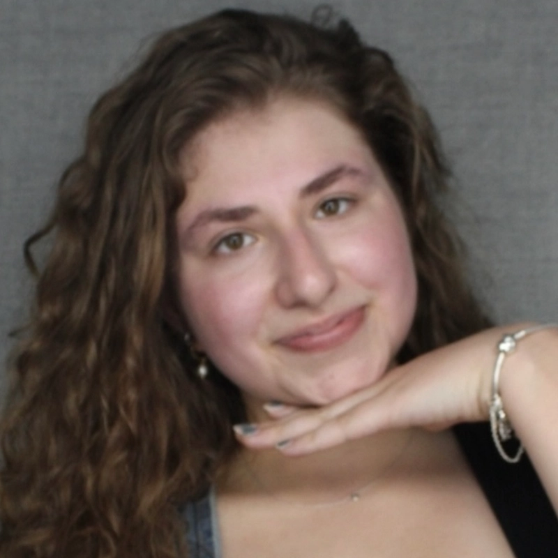 A young woman with long wavy brown hair smiles softly, resting her chin on her hand. She wears a silver bracelet and a dark top, with a gray background behind her. Her expression is relaxed and friendly, reminiscent of the calm presence often captured in portraits by Asher Alcantara.