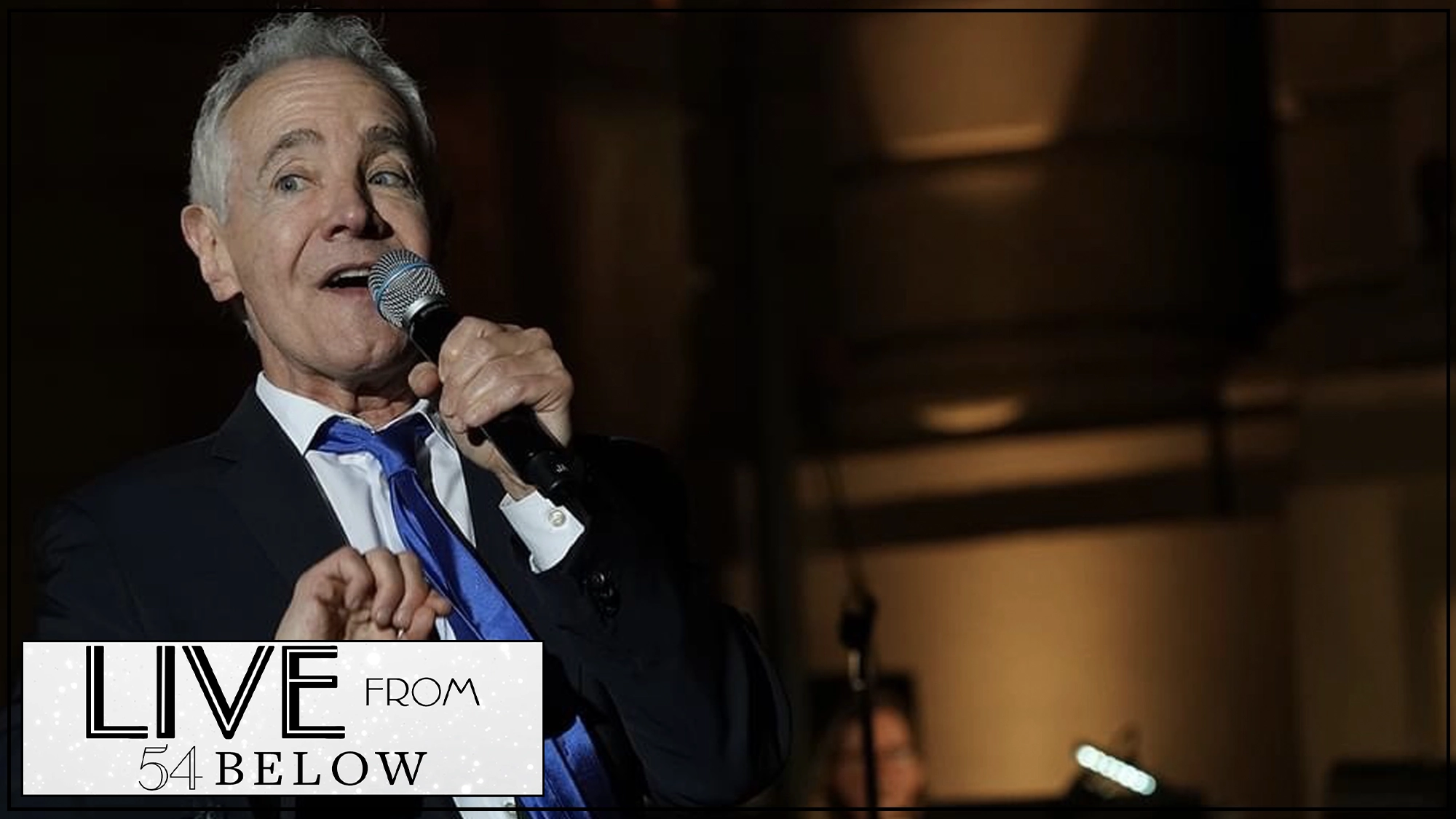 LIVE Jason Graae Show Page A gray-haired man in a suit and blue tie sings into a microphone onstage. He gestures with one hand, looking animated. Warm stage lighting and a shadowy background set the scene as Jason Graae delivers a captivating singing performance.