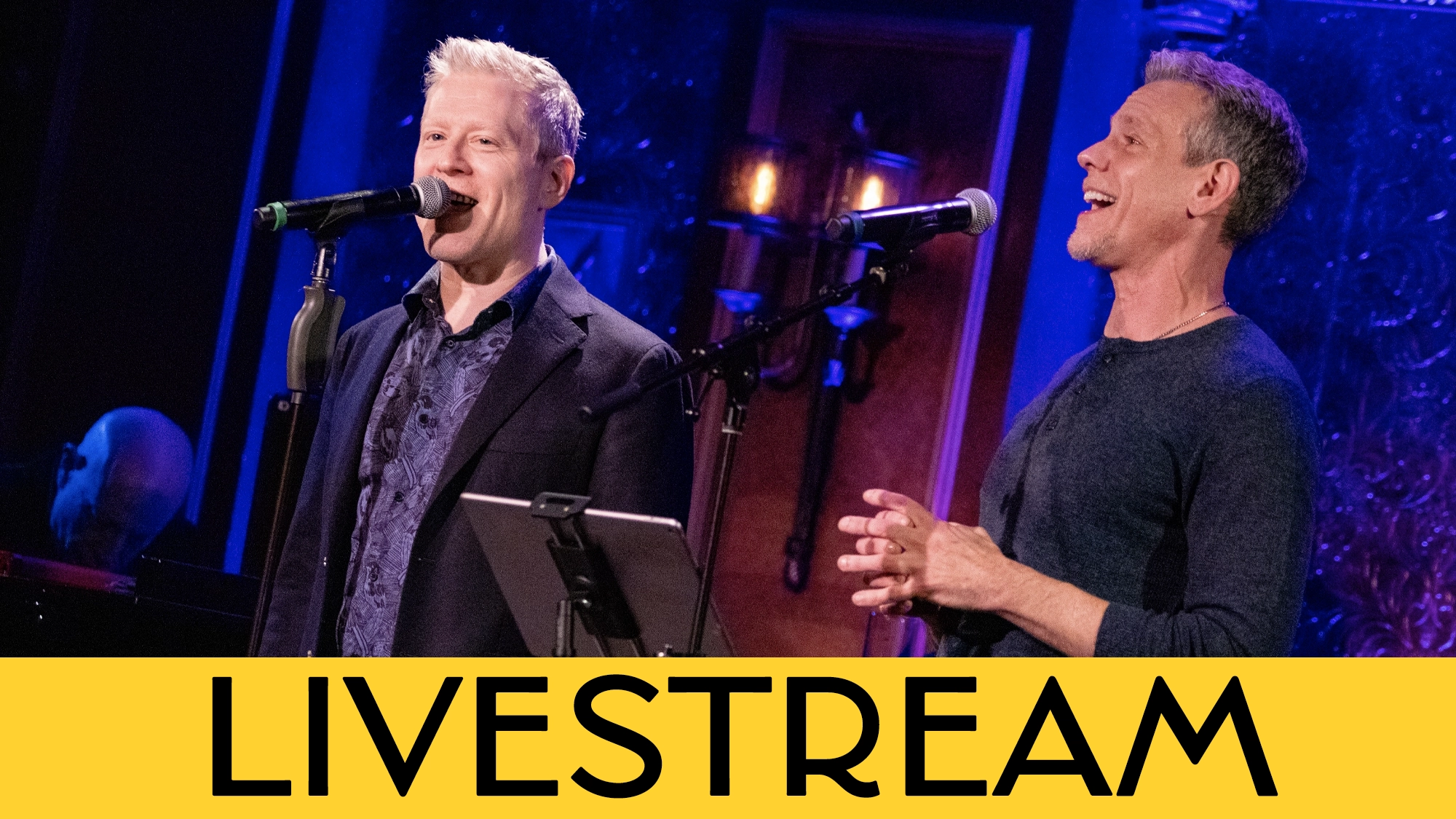Two men, Adam Pascal and Anthony Rapp, sing into microphones on a dimly lit stage with ornate decor. One wears a dark suit, the other a dark shirt. Sheet music is on a stand between them. The lower part of the image features a yellow banner with the word “LIVESTREAM” in bold black letters—a captivating moment from their 