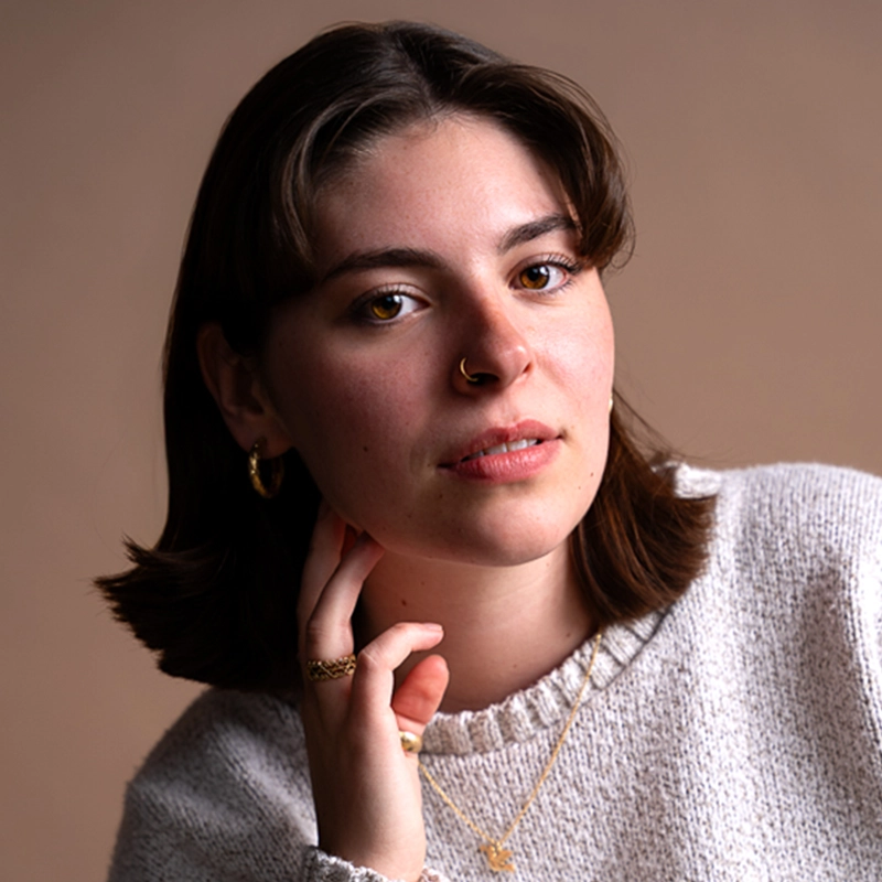 A young woman with straight brown hair and light skin wears a cream sweater, gold hoop earrings, nose ring, and gold necklace. Channeling effortless 