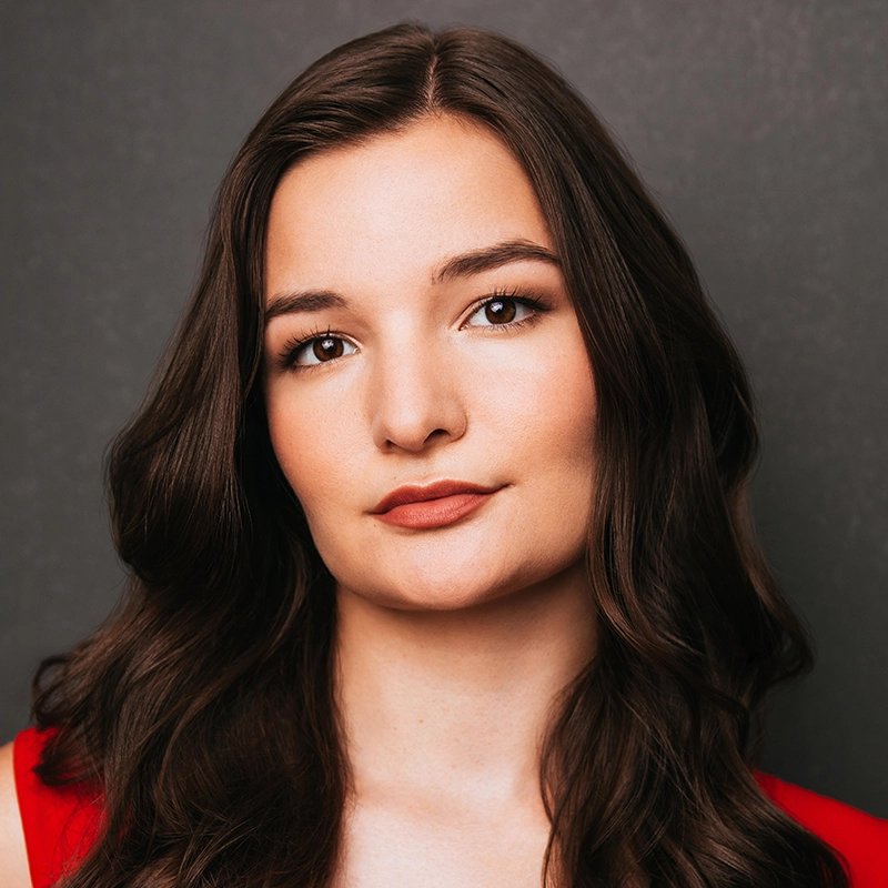 A young woman with long, wavy brown hair and fair skin looks at the camera with a calm expression. She wears a red top and has subtle makeup. The background is dark gray and plain, drawing focus to Casey Keeler’s poised presence.