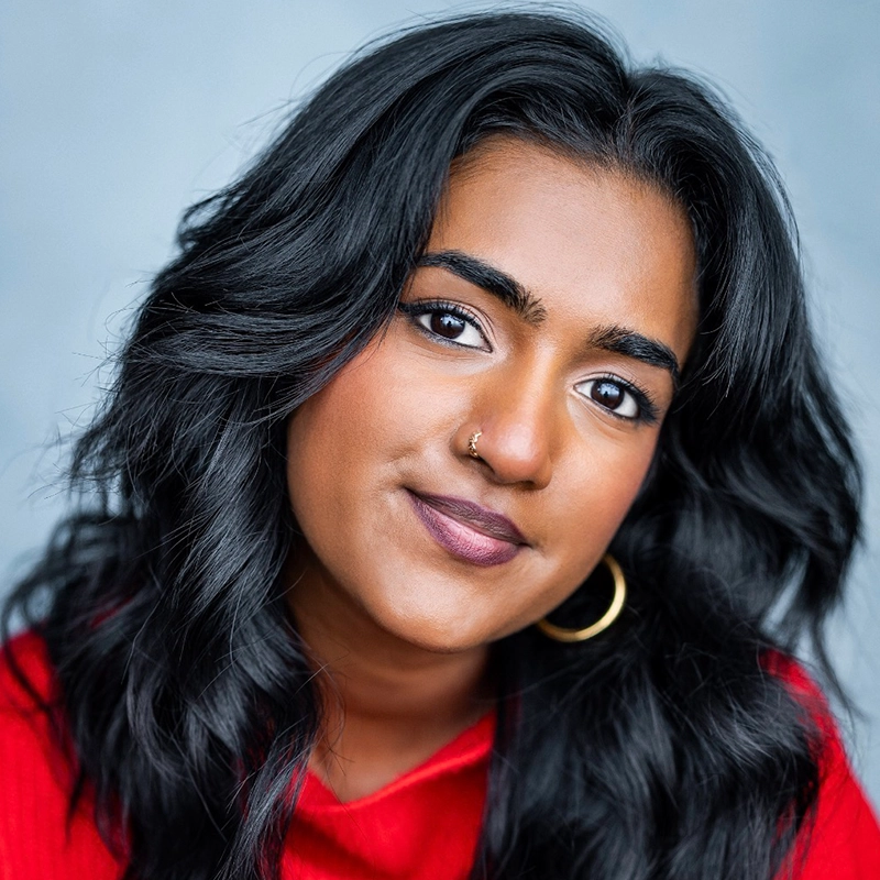 A woman with wavy black hair, wearing a red top, gold hoop earrings, and a nose ring, looks at the camera with a soft smile against a muted blue background—an image that could easily capture Alexandra Zografos's understated elegance.