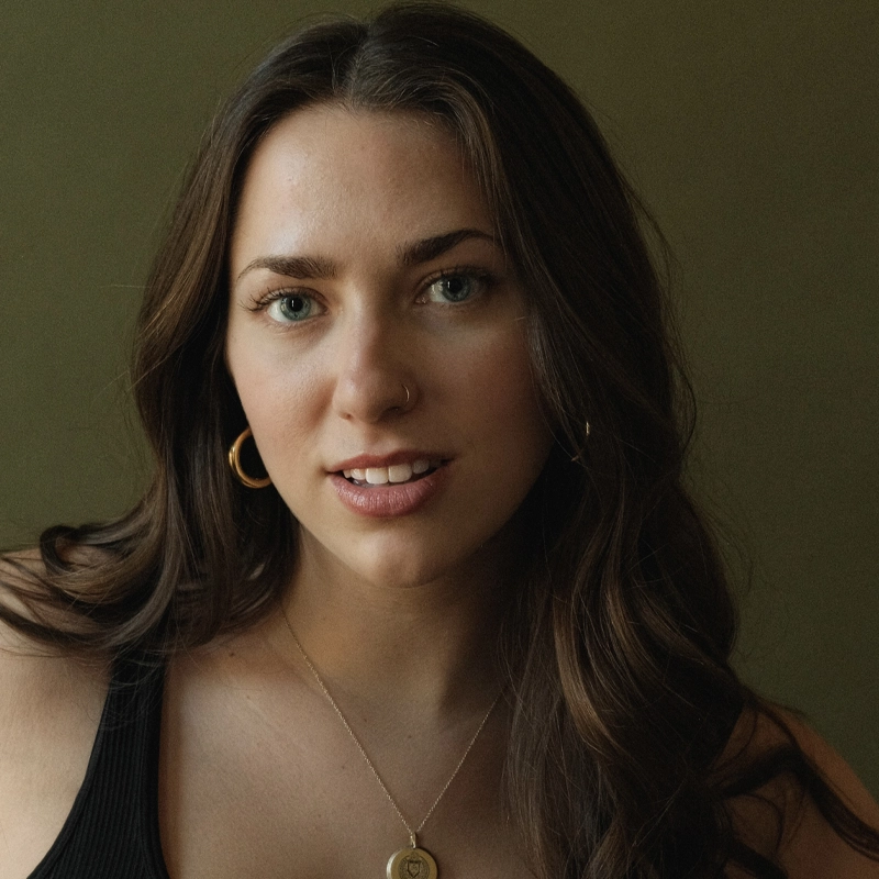A woman with long brown hair and blue eyes looks at the camera, smiling softly. She wears gold hoop earrings, a thin necklace with a round pendant, and a black tank top. The background is plain and olive green. The lighting is soft and natural.