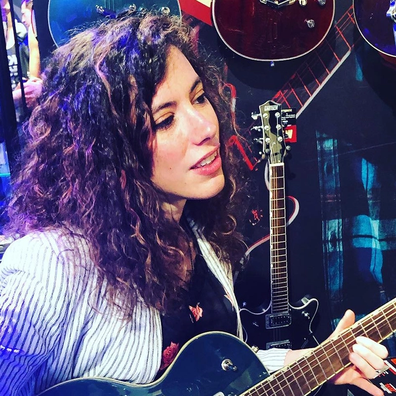 Gabby Sherba, a woman with curly hair and a striped jacket, strums an electric guitar. She is surrounded by guitars displayed on a wall, and colorful lighting highlights her face. She appears focused and engaged with the instrument.