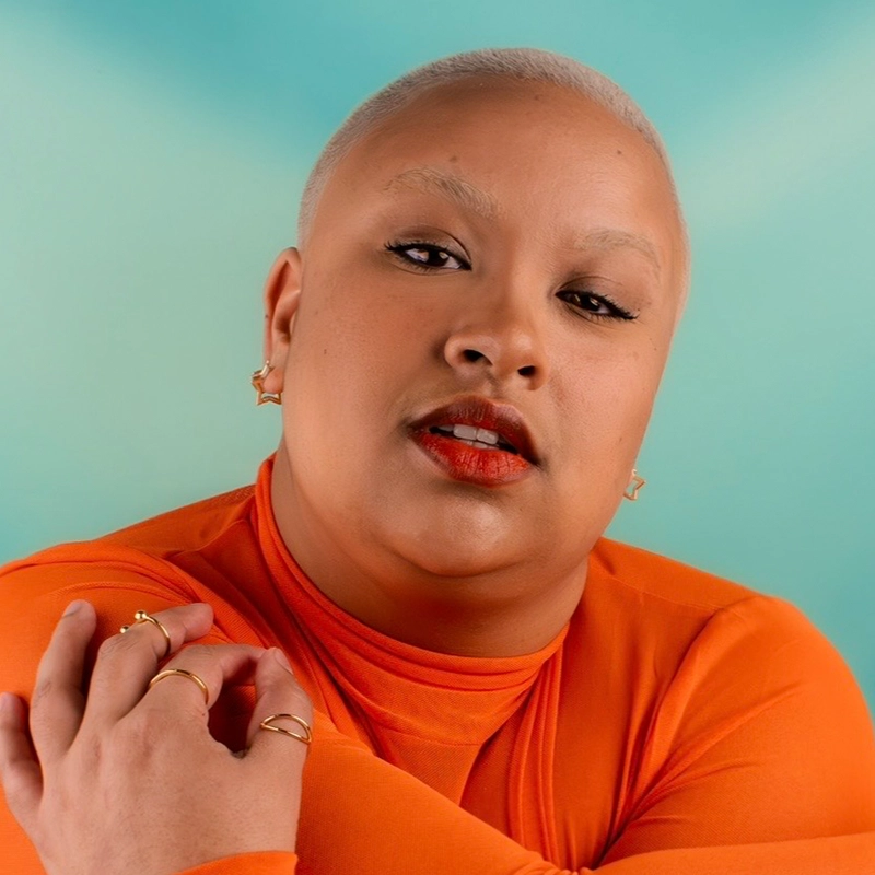 Gabrielle Farley, with very short, bleached hair and bleached eyebrows, poses confidently against a light blue background. She wears an orange top, gold rings, and star earrings, complemented by bold red lipstick. Her arms are crossed as she gazes assuredly at the camera.