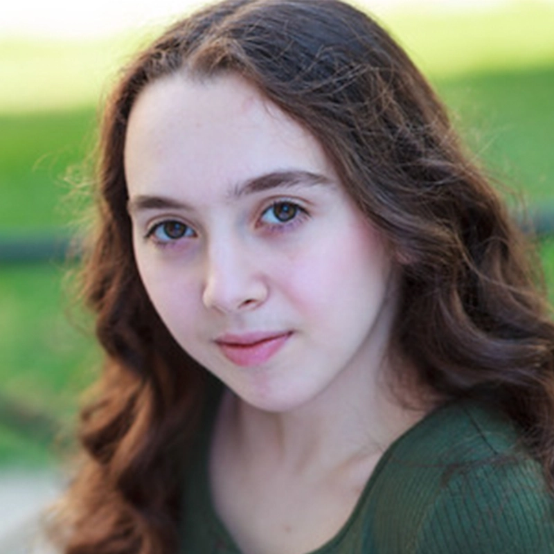 A young woman with long, wavy brown hair and fair skin looks directly at the camera. She has light hazel eyes and is wearing a green top. The background is softly blurred greenery, suggesting an outdoor setting on a sunny day. This portrait captures Michelle Zink-Muñoz in a moment of natural beauty and quiet confidence.