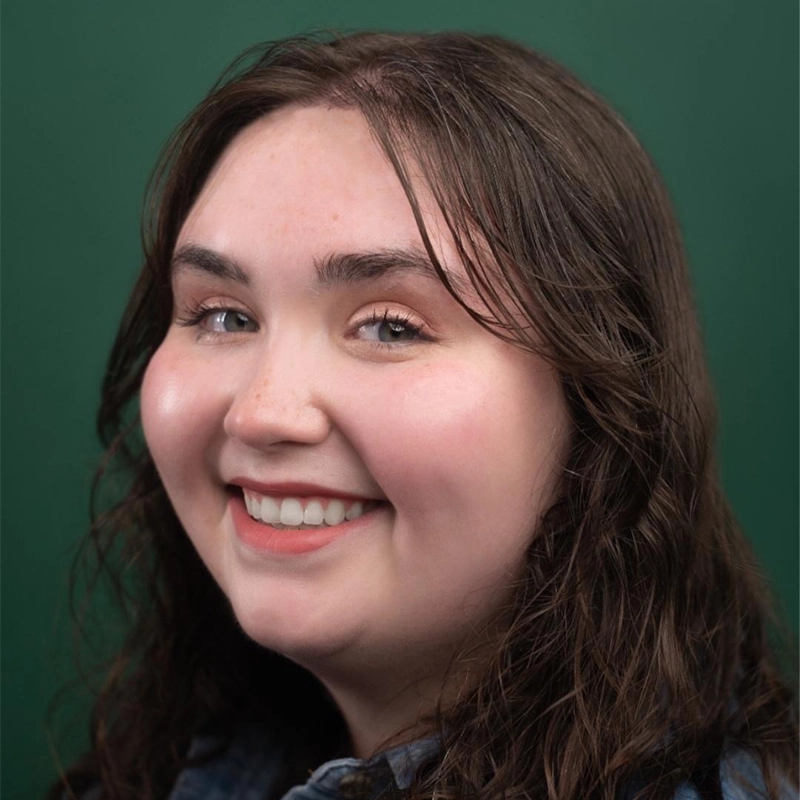 A young woman with long, wavy brown hair smiles warmly at the camera. She has fair skin, full cheeks, and is wearing a denim shirt. The background is a solid dark green. Her expression is friendly and relaxed, perfectly capturing the approachable vibe of someone you might hear say, 