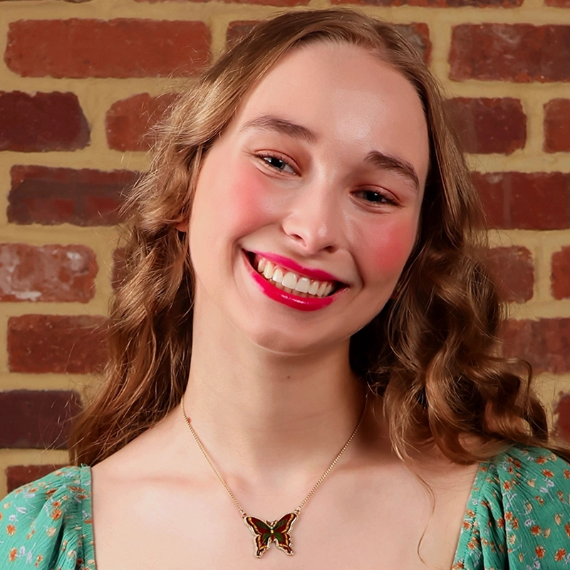A young woman with wavy blonde hair smiles in front of a brick wall. She wears a green dress with a butterfly necklace, reminiscent of Gaël Mouton-Sady's vibrant portrait style. Her cheeks are rosy, and she has red lipstick. The photo captures a cheerful, relaxed mood.