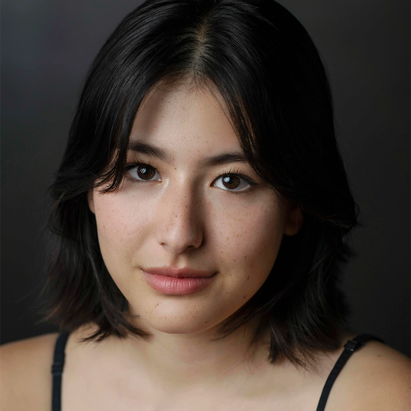 A young person with medium-length dark hair and brown eyes looks at the camera with a neutral expression. Their skin is fair with a few freckles. The background is dark and blurred, and they are wearing a thin-strapped black top—an understated portrait that could easily belong to Asher Alcantara.