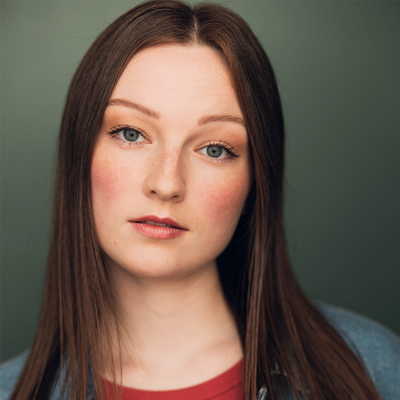A young woman with straight brown hair poses against a green background. She has light skin, blue eyes, and faint freckles. Casey Keeler wears natural makeup, a denim jacket, and a red top, looking directly at the camera with a neutral expression.