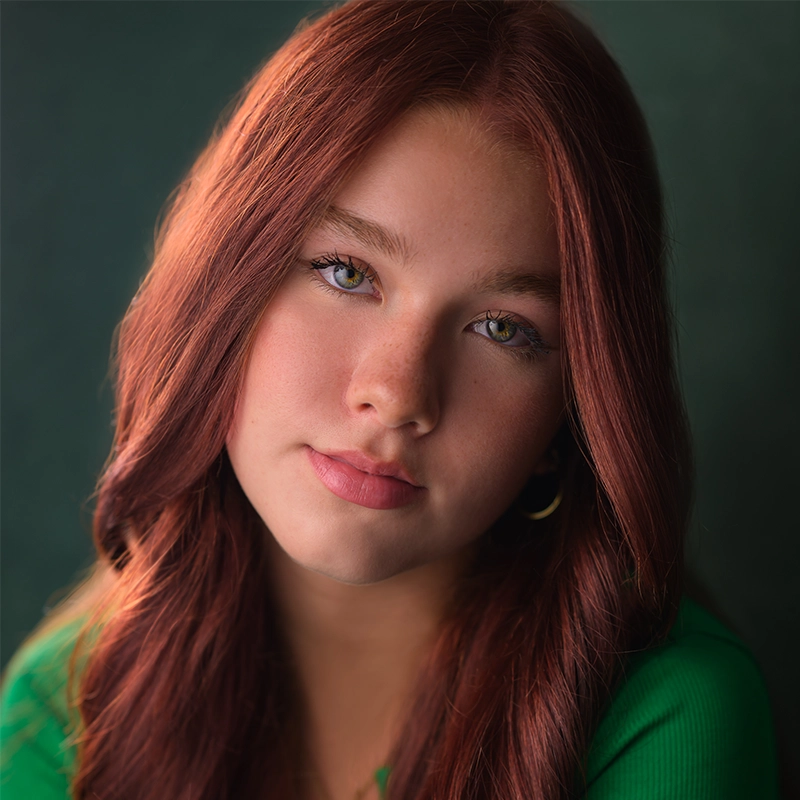 A young woman with long, wavy auburn hair and light eyes looks at the camera. She wears a green top and gold hoop earrings. Her expression is calm and thoughtful, with soft lighting highlighting her features against a dark green background. The portrait captures Millie Yocum’s quiet confidence, her presence almost luminous as she surfaces gently from the shadows.