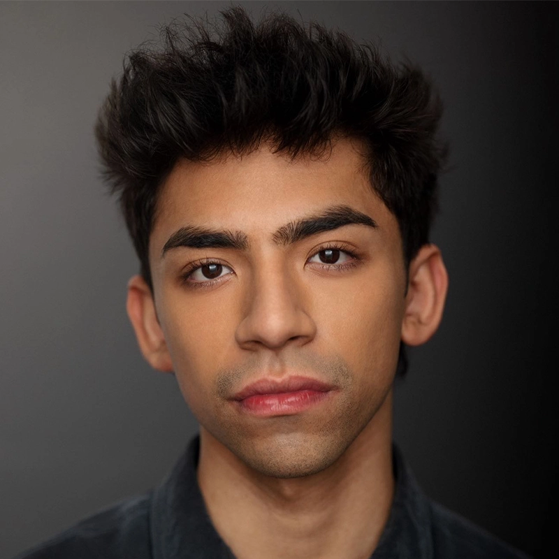 A young man with short, dark, tousled hair and thick eyebrows stares directly into the camera with a neutral expression. He has medium-toned skin and wears a dark shirt. The background is a smooth gradient from dark gray to black, providing a subtle yet striking frame for Saylor Tucker’s composed presence.