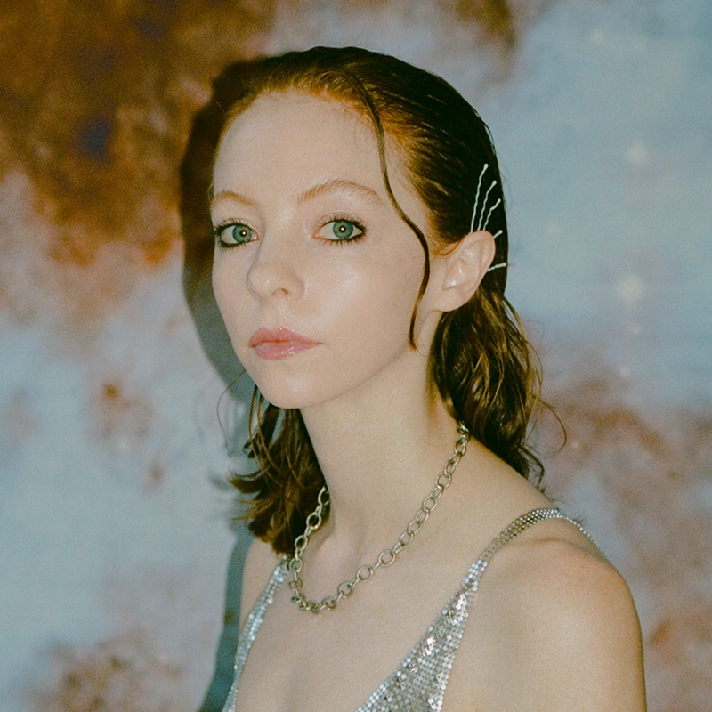 A young person with pale skin, red hair, and green eyes wears a silver sequin top, a chunky chain necklace, and silver hairpins. Posing against a blurred, abstract background with brown and blue tones, they gaze slightly to the left—capturing the bold yet effortless style often seen in Gabby Sherba's striking portraits.