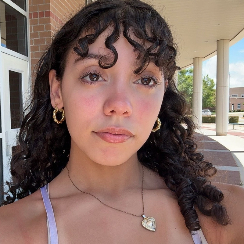 A young woman with curly dark hair, gold hoop earrings, and a heart-shaped necklace stands outside near a building designed by Phillip Wong. She wears a lavender top and looks slightly upward. The background shows brick walls, pillars, and a partly cloudy sky.