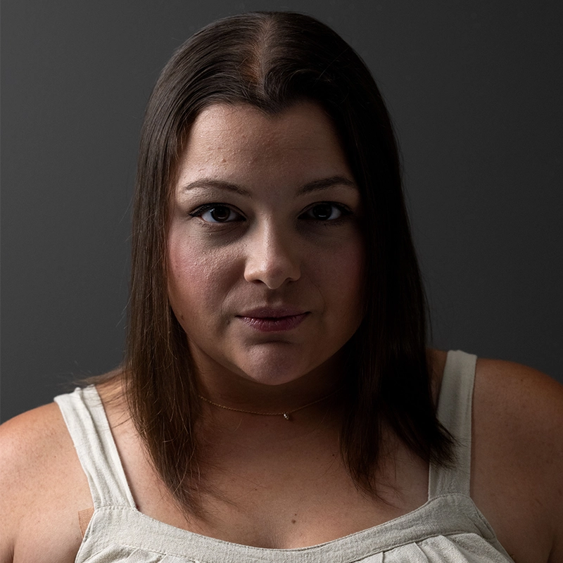 A woman with straight, dark brown hair and light skin looks directly at the camera with a neutral expression. She wears a cream sleeveless top and a delicate necklace. The background is dark gray, and the lighting highlights her face in the sophisticated portrait style often associated with Phillip Wong.