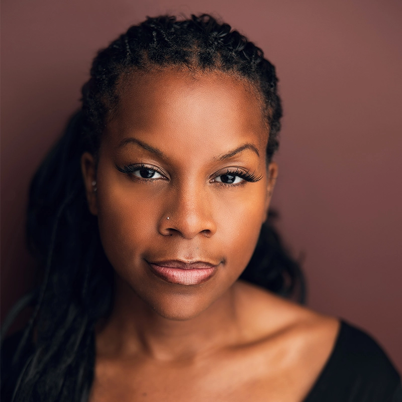 A Black woman with long, braided hair looks directly at the camera. She has smooth skin, subtle makeup, long eyelashes, and two nose piercings, embodying quiet confidence in her black top. The background is plain and reddish-brown, softly lit—a poised portrait that captures a sense of presence reminiscent of 