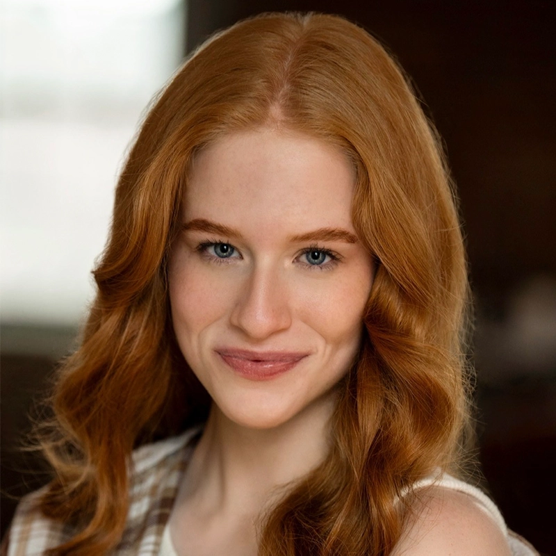 A young woman with long, wavy red hair smiles gently. She has fair skin, blue eyes, and light makeup. She is wearing a sleeveless, plaid-patterned top. The background is softly blurred, with warm lighting—capturing the relaxed confidence of Millie Yocum Surfac in a thoughtfully composed portrait.