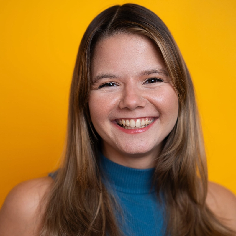 A young woman with long, light brown hair smiles warmly at the camera. She wears a sleeveless blue mock-neck top. The background is a solid, vibrant yellow, adding brightness and contrast to the image. The lighting highlights her cheerful expression.