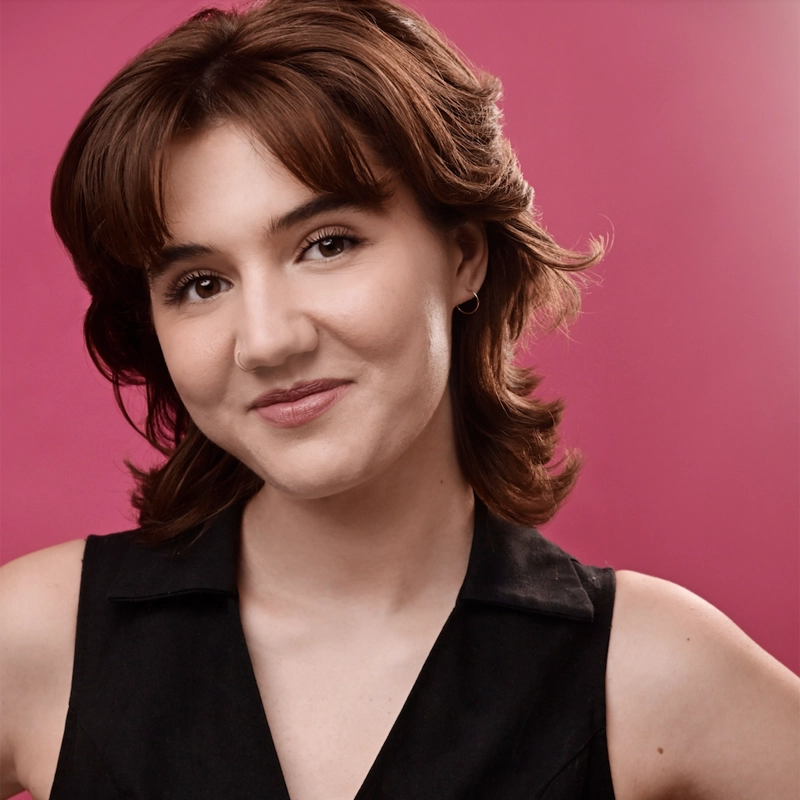 Lauren Manna, a young woman with short, wavy brown hair, poses against a pink background. She smiles softly, wearing a black sleeveless top, a small nose ring, and a hoop earring. Her expression is calm and confident, with her arms slightly visible in the frame.
