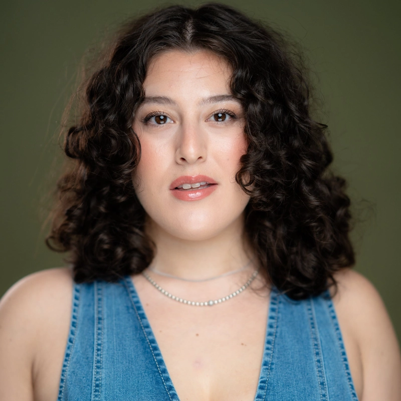 Giovanna Marchese, a woman with shoulder-length, dark curly hair and light skin, faces forward. She wears light makeup and a sleeveless blue denim top with a deep V-neck. Her expression is neutral, with lips slightly parted. The background is plain olive green.