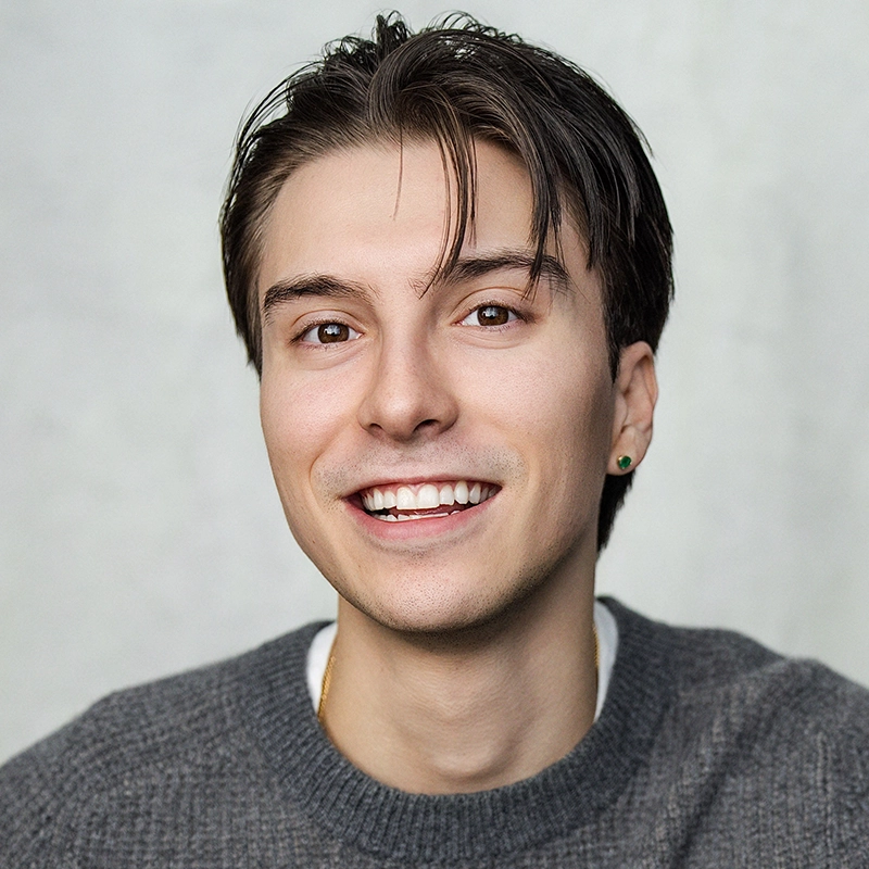 Young person with fair skin, straight brown hair parted to the side, and brown eyes smiles at the camera. They wear a gray knit sweater, a small earring, and sit in front of a light, neutral background. The mood is friendly and casual, capturing the approachable presence of Asher Alcantara.