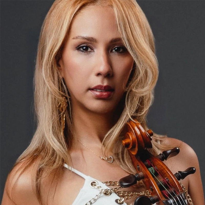 Maria G. Figueroa, a woman with long, light blonde hair wearing a white top and gold jewelry, holds a violin upright. She has a calm expression and stands against a plain dark background. The violin’s scroll and pegs are visible near her shoulder.