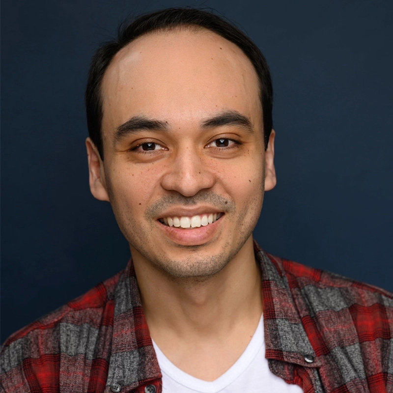 A man with short black hair and light skin smiles at the camera. He is wearing a red and gray plaid shirt over a white t-shirt. The background is a solid dark blue.