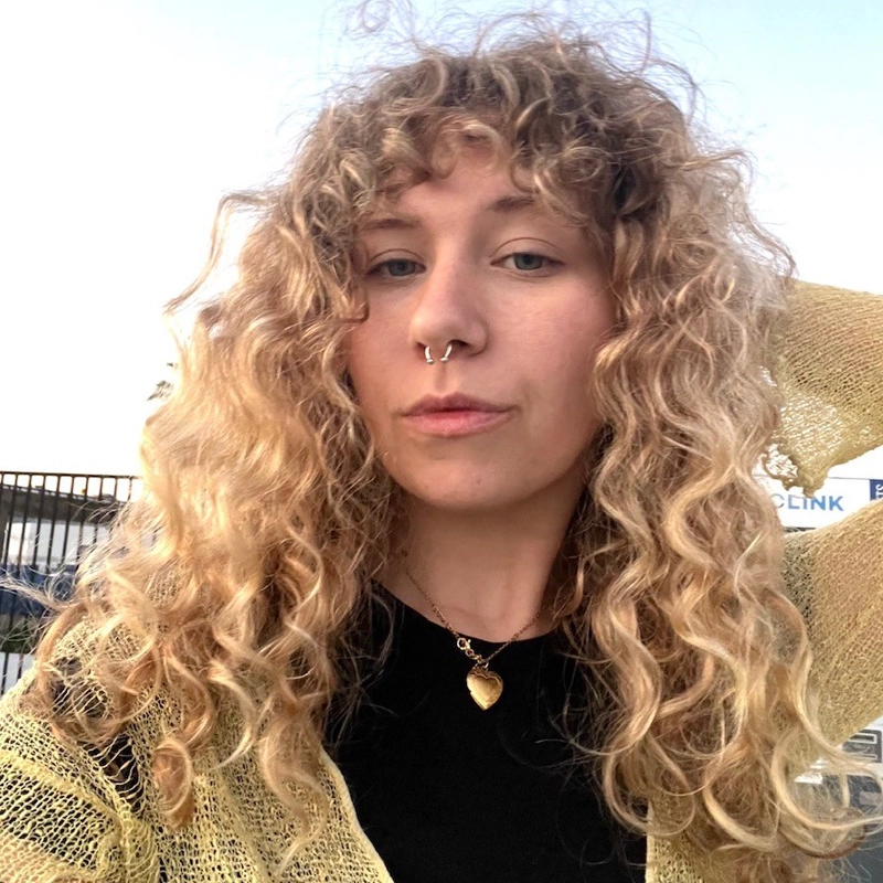 A person with curly blonde hair reminiscent of Honey Marmalade, a gold septum ring, and a heart necklace poses outdoors at sunset, wearing a black top and a shimmery gold shawl. The background shows a fence and a partially visible sign, capturing the effortless style of Olivia Klein.