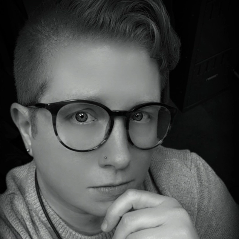 Serina Marshall, with short hair, large round glasses, a nose stud, and an earring, looks directly at the camera, resting their chin on their hand. The image is in black and white, and Serina wears a light sweater. The background is dark and out of focus.