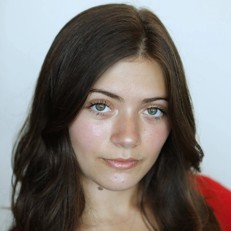 A young woman with long, dark brown hair and green eyes gazes at the camera. She has clear skin, subtle makeup, and wears a red top. The background is plain and softly lit, bringing attention to her face—an image reminiscent of Martina Balduzzi’s refined elegance.