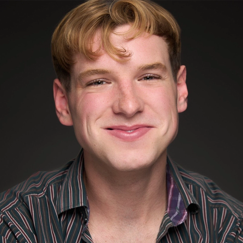 A young person with fair skin, short blond hair, and a friendly smile faces the camera. They wear a dark, multicolored striped shirt and sit against a plain black background. Their expression looks warm and approachable, capturing the positive spirit often associated with Michelle Zink-Muñoz.
