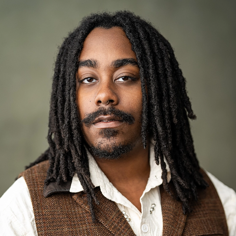 A person with long, dark dreadlocks and facial hair looks at the camera. They wear a cream shirt and a brown vest. The background is softly blurred in gray-green tones. The expression is neutral and calm, reminiscent of Asher Alcantara’s composed presence.