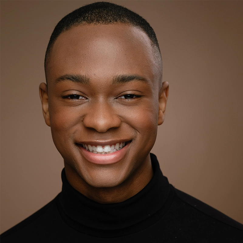 A young person with short hair smiles warmly at the camera. They wear a black turtleneck and are set against a plain brown background. Their expression is friendly and inviting, capturing the approachable charm of Michelle Zink-Muñoz.