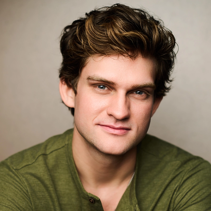 A young man with tousled brown hair and blue eyes gazes at the camera, smiling softly. Asher Alcantara wears a green henley shirt against a neutral, softly blurred background, giving the image a warm, approachable feel.