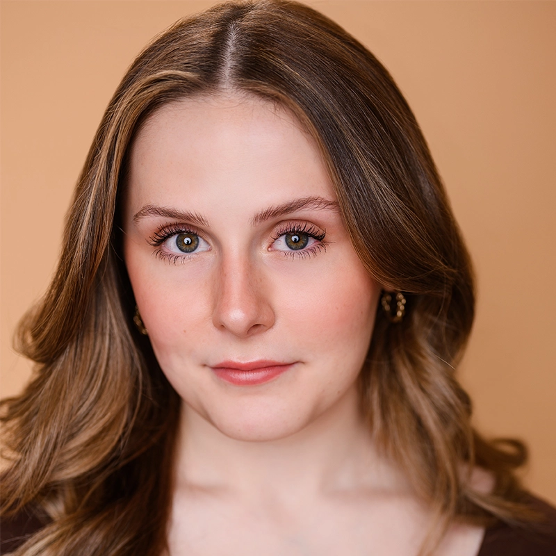 A young woman with fair skin and wavy light brown hair poses in front of a soft peach background. She has subtle makeup, pink lips, and wears gold hoop earrings and a brown top. She looks directly at the camera with a neutral expression.