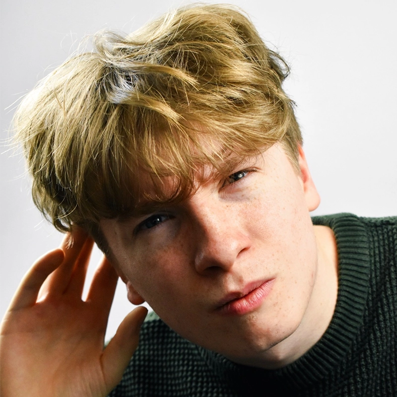 Young person with tousled blonde hair, wearing a green knit sweater, leans forward and cups one ear, squinting slightly, as if trying to hear or understand something—an expression reminiscent of Gaël Mouton-Sady’s curious and intent demeanor. Background is plain and light-colored.